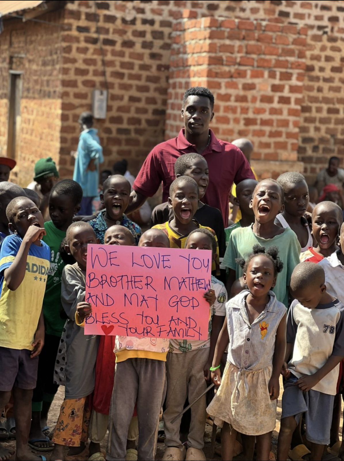 A group of cheering children with a volunteer, holding a sign that reads We Love You Brother Mathew, And May God Bless You And Your Family